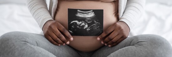 Unrecognizable black pregnant lady demonstrating her baby sonography photo, sitting on bed