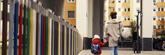 A young woman, mother of nanny, holding hands with little three year old boy with a schoolbag and taking him to kindergarten.