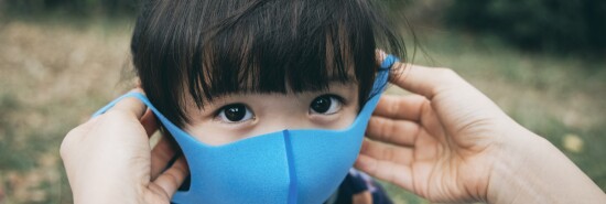 Mother putting on surgical mask for little daughter in the park to prevent the spread of cold and flu and viruses