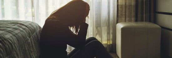 A depressed woman sits in a dark bedroom with her hands covering her face.